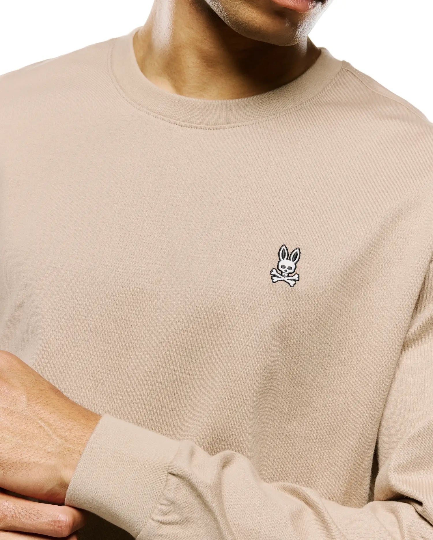Mens Norman Long Sleeve Relaxed Fit Tee in beige, featuring a soft jersey knit texture and a small, embroidered black and white Psycho Bunny skull logo on the left chest.