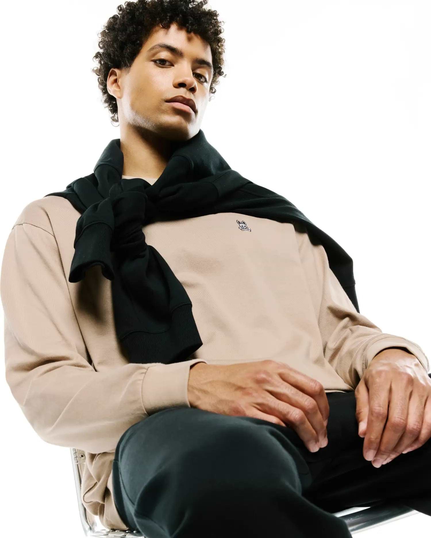 Mens Norman Long Sleeve Relaxed Fit Tee in beige jersey, featuring a small embroidered black Psycho Bunny skull logo on the left chest, paired with black pants and a black sweater tied over the shoulders.