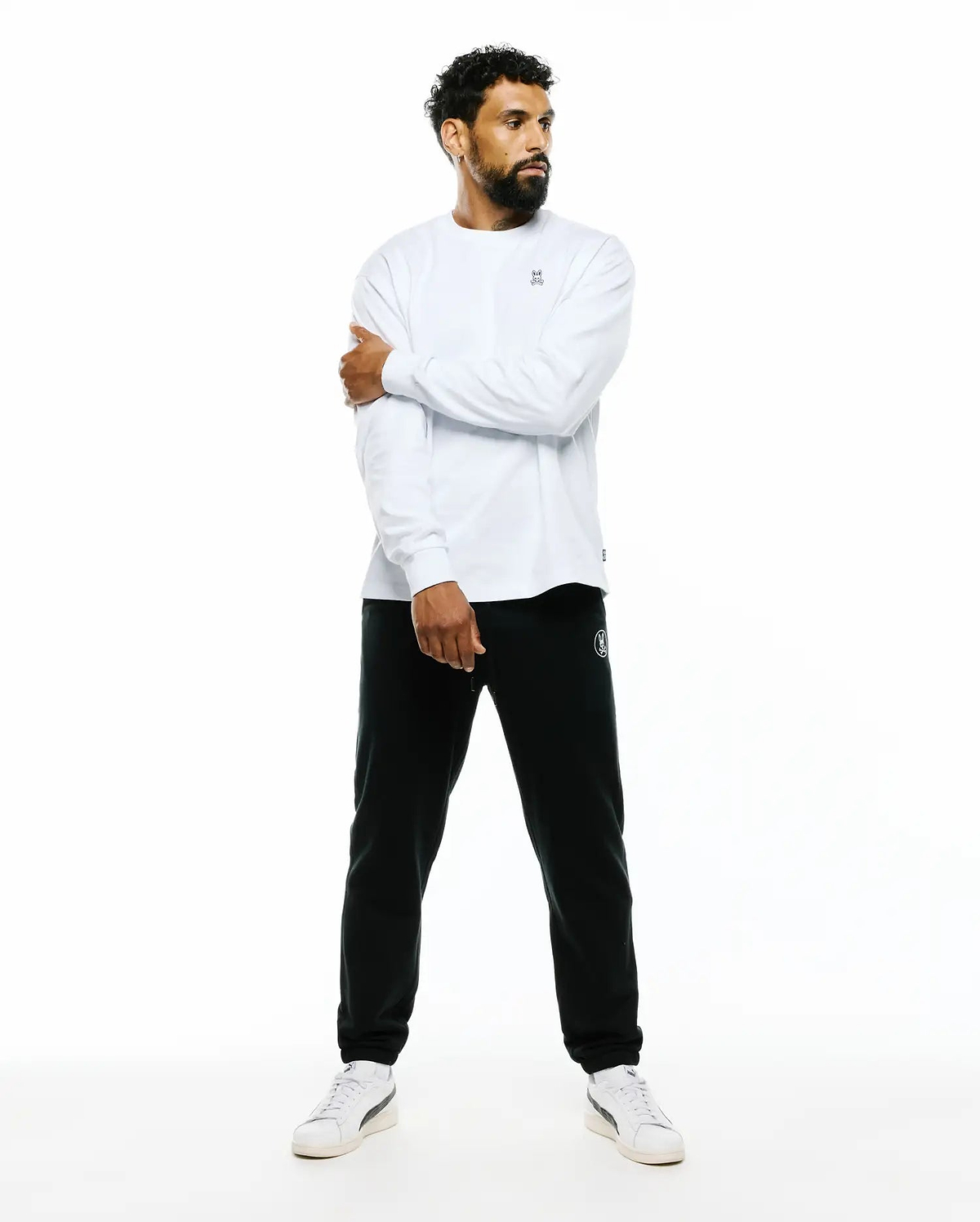 Mens Norman Long Sleeve Oversized Fit Tee in white jersey knit with a small embroidered Psycho Bunny skull logo on the chest, paired with black French terry joggers featuring a white circular bunny logo on the left thigh.