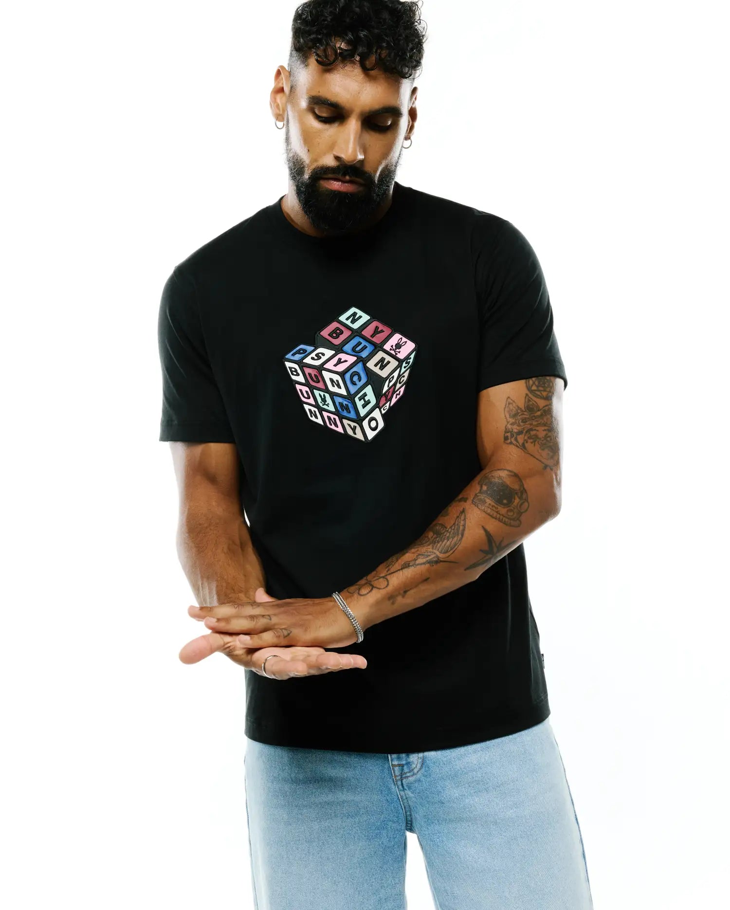 Mens Eddy Graphic Tee, a black classic fit jersey t-shirt featuring a large multicolored Rubik's Cube-style graphic on the chest spelling PSYCHO BUNNY with the iconic skull logo, and a small woven tag on the hem.