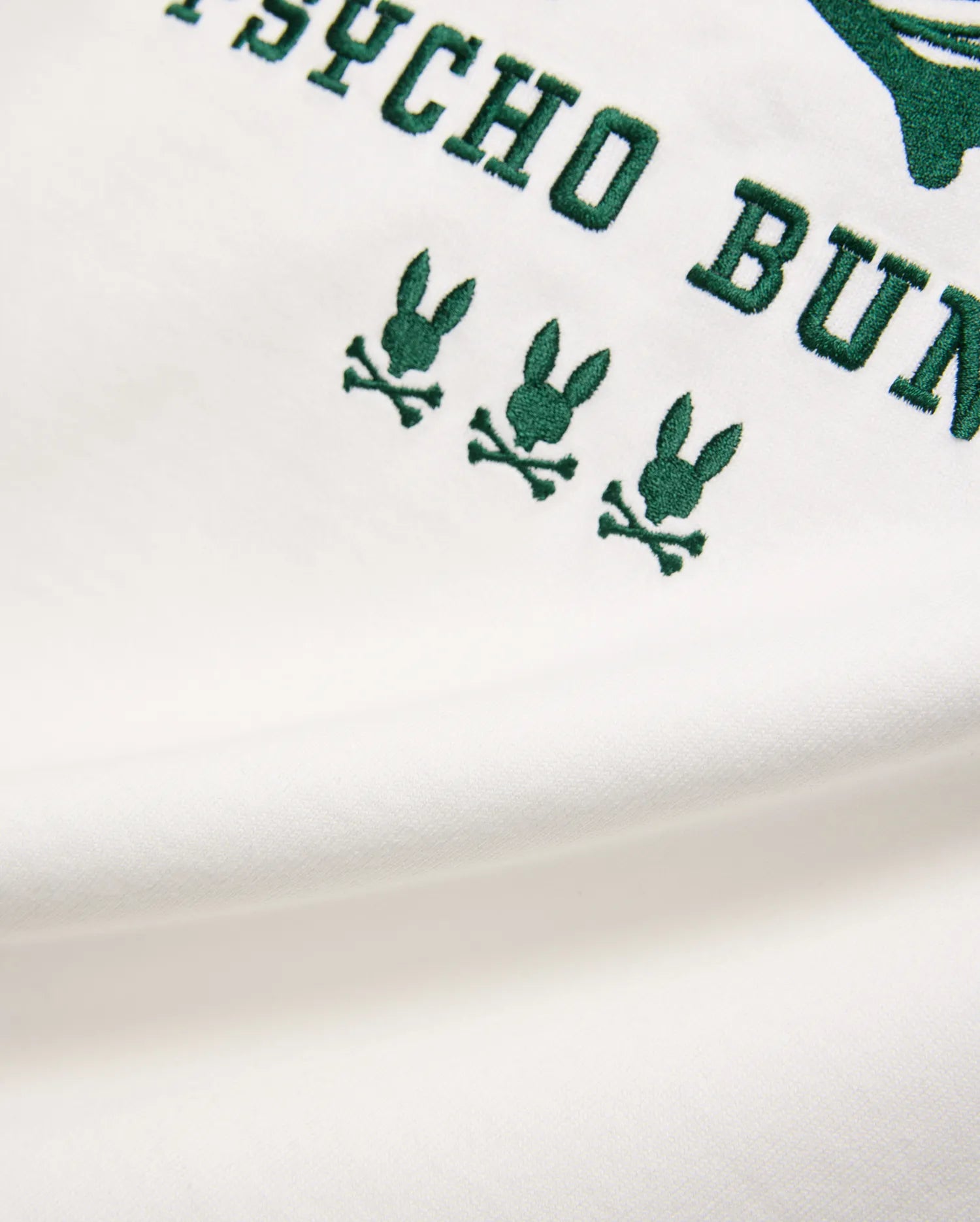 Mens Mike Graphic Jersey Tee, a premium white jersey knit fabric with bold dark green embroidered Psycho Bunny text and three distinct, small bunny skull logos, showcasing refined streetwear style.