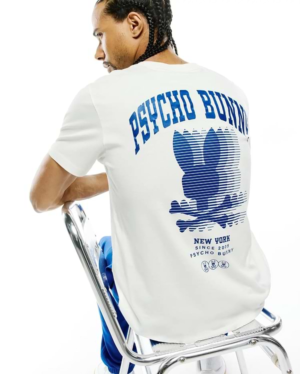 Mens Nathaniel Back Graphic Tee, a classic fit white jersey t-shirt featuring a large blue Psycho Bunny text and striped bunny skull logo graphic on the back, with New York Since 2005 branding.