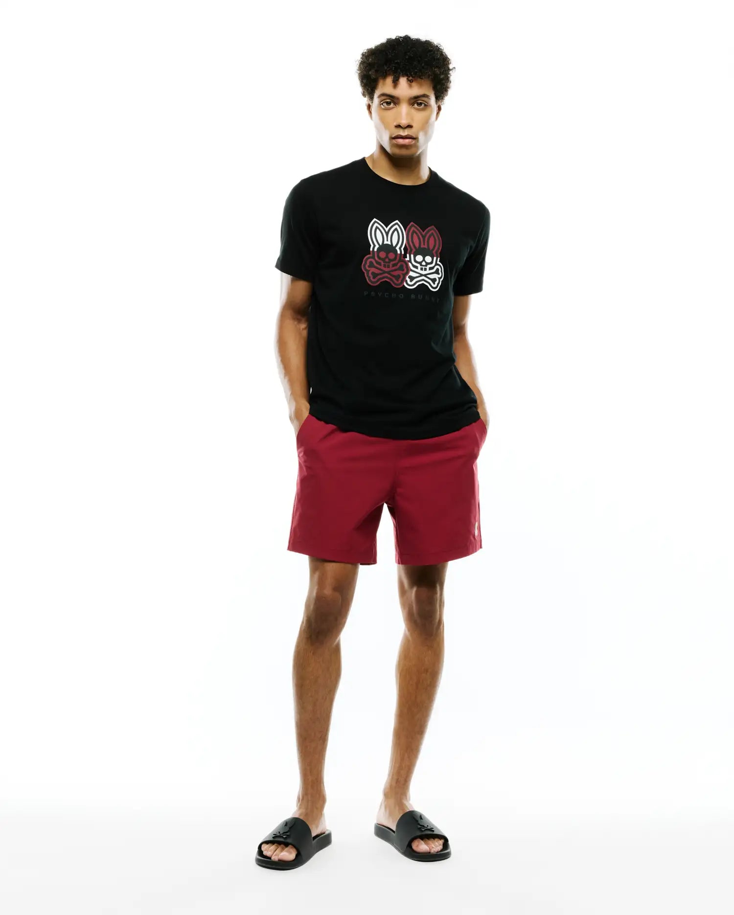 Mens Ledger Graphic Tee, a black jersey t-shirt with a large white and red Psycho Bunny skull graphic on the chest, paired with red shorts featuring a small embroidered bunny logo.