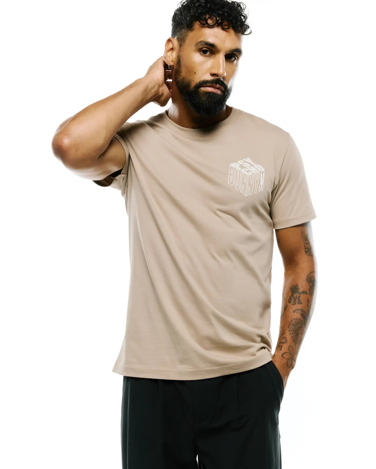 Mens Conan Back Graphic Tee, a classic fit light tan jersey knit t-shirt with a prominent white 3D stacked Psycho Bunny text and skull logo graphic on the left chest.