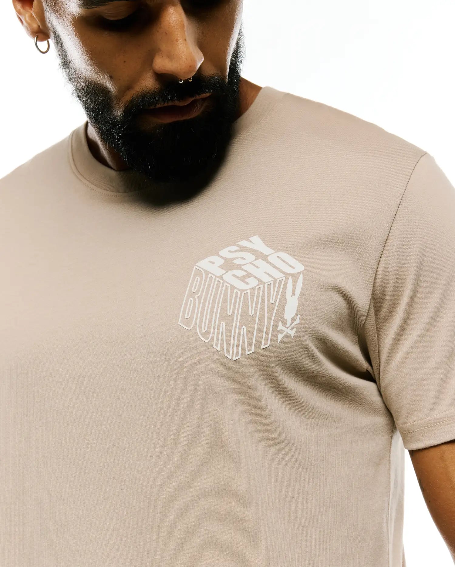 Mens Conan Back Graphic Tee, a classic fit light beige jersey t-shirt with a prominent white 3D block Psycho Bunny text graphic and skull logo on the chest.
