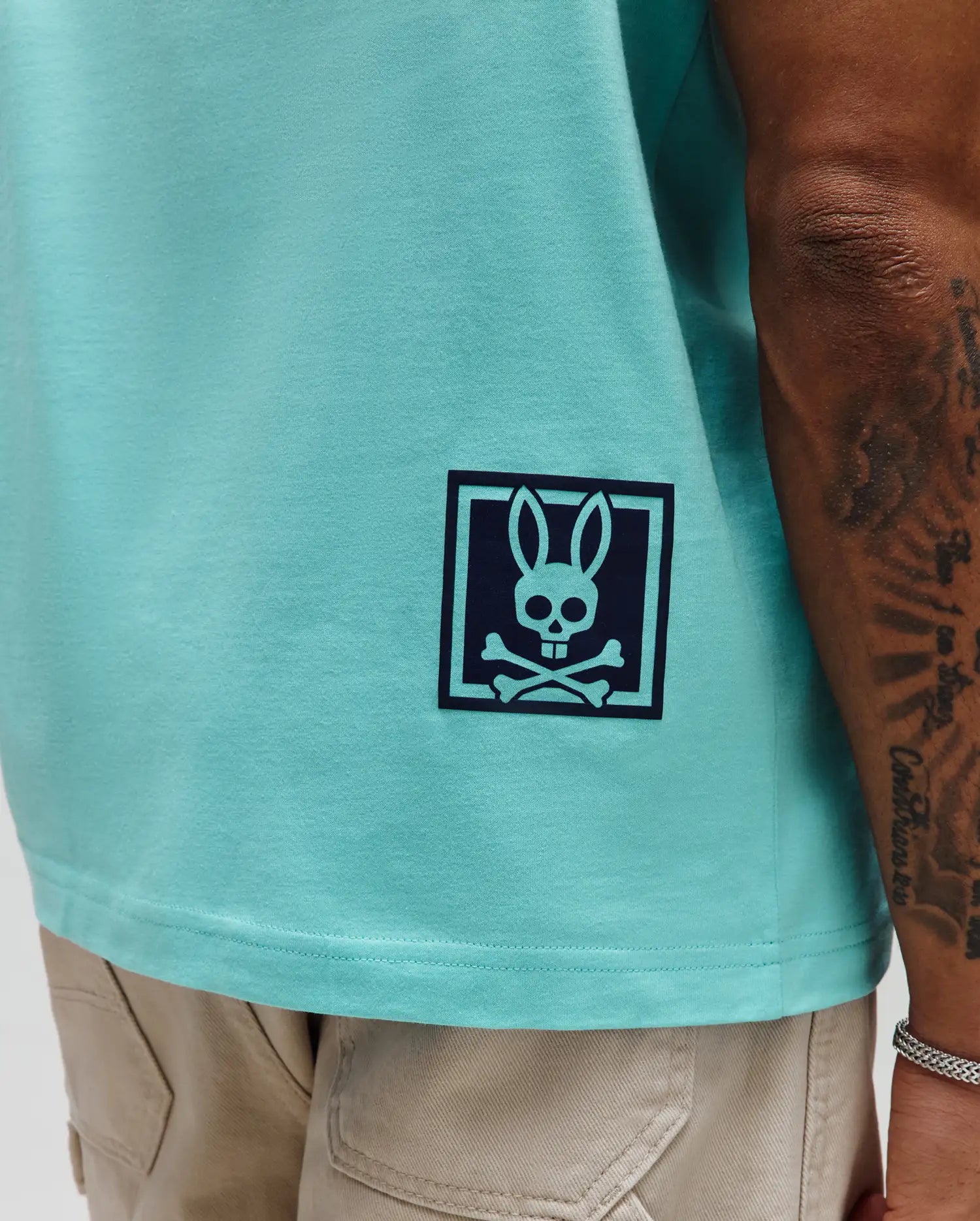Mens Keon Graphic Tee, aqua smooth jersey knit, classic fit, featuring a large navy Psycho Bunny skull and crossbones logo in a square frame on the lower left hem.