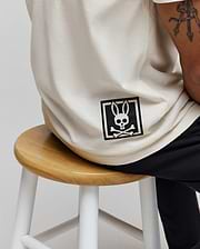 Mens Keon Graphic Tee in cream, featuring a relaxed fit and smooth jersey knit. A large black Psycho Bunny skull and crossbones logo graphic is printed on the lower back, framed by a square.