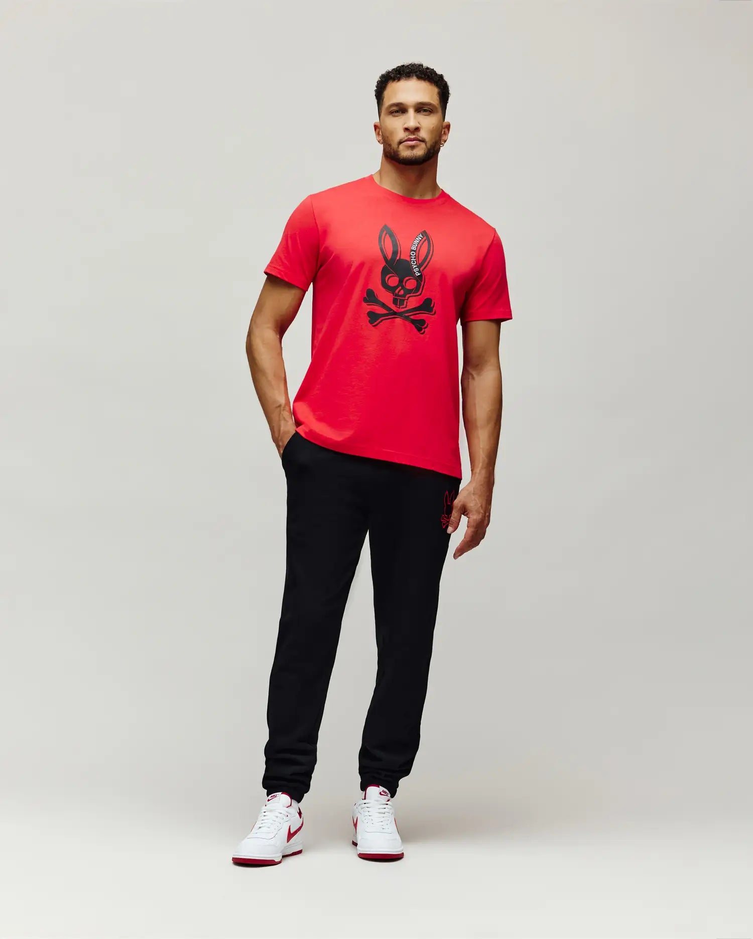 Mens Serge Embroidered Bunny Graphic Tee: Vibrant red classic fit jersey t-shirt featuring a large central black Psycho Bunny skull and crossbones graphic with vertical brand text on the ear.