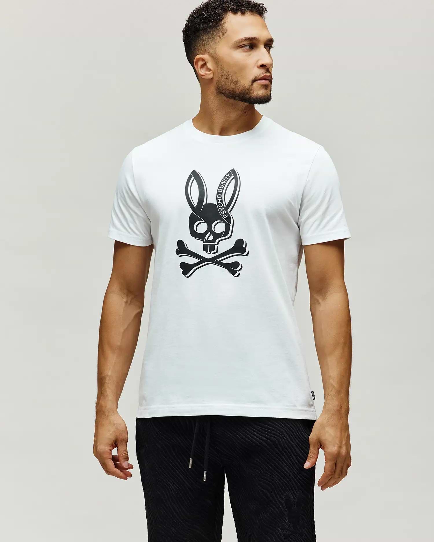 Mens Serge Embroidered Bunny Graphic Tee, a classic fit white jersey knit t-shirt with a large black Psycho Bunny skull and crossbones chest graphic, featuring PSYCHO BUNNY text in the ears, and a small woven brand tag on the left hem.