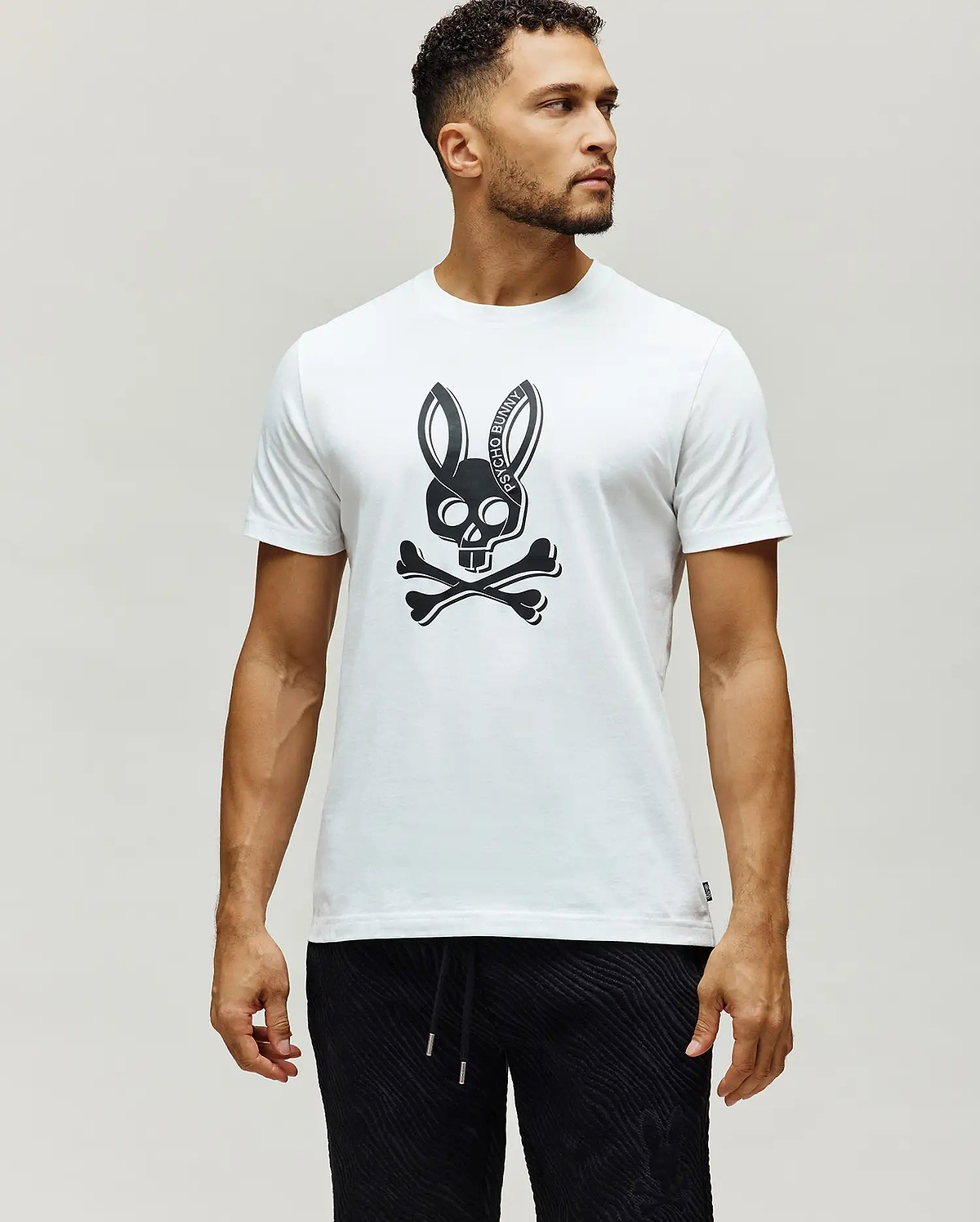 Mens Serge Embroidered Bunny Graphic Tee, a classic fit white jersey knit t-shirt with a large black Psycho Bunny skull and crossbones chest graphic, featuring PSYCHO BUNNY text in the ears, and a small woven brand tag on the left hem.