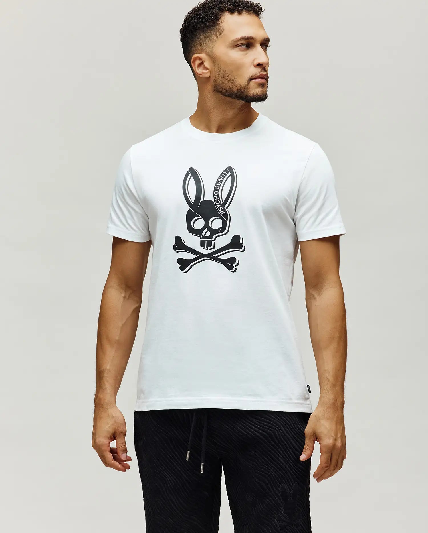 Mens Serge Embroidered Bunny Graphic Tee, a classic fit white jersey knit t-shirt with a large black Psycho Bunny skull and crossbones chest graphic, featuring PSYCHO BUNNY text in the ears, and a small woven brand tag on the left hem.