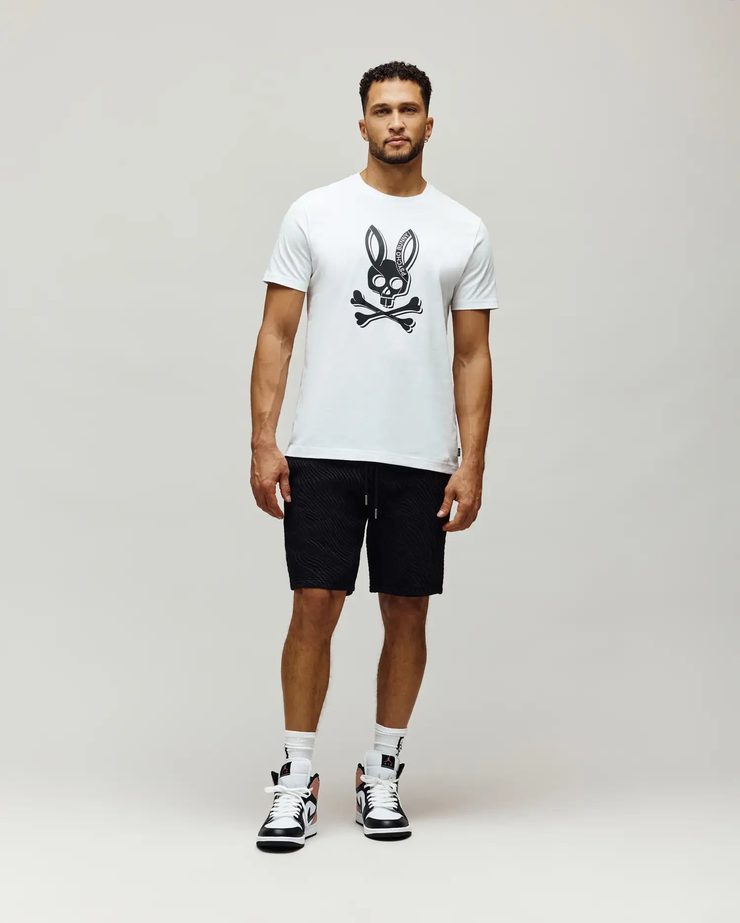Mens Serge Embroidered Bunny Graphic Tee in white, featuring a large black Psycho Bunny skull graphic on the chest. Paired with black textured drawstring shorts.