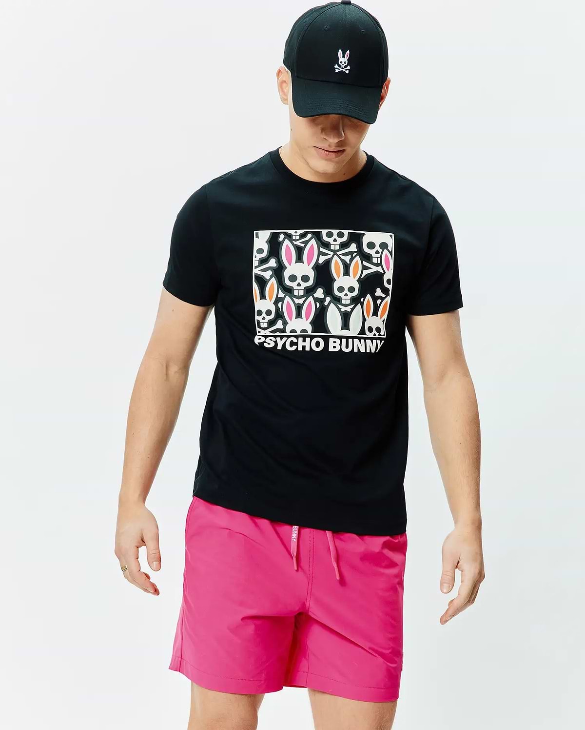 Mens Byron Graphic Tee, black jersey knit, with a large central graphic of colorful Psycho Bunny skulls and bones. Paired with bright pink drawstring shorts and a black baseball cap featuring an embroidered pink-eared Psycho Bunny logo. (4)