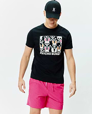 Mens Byron Graphic Tee, black jersey knit, with a large central graphic of colorful Psycho Bunny skulls and bones. Paired with bright pink drawstring shorts and a black baseball cap featuring an embroidered pink-eared Psycho Bunny logo. (4)