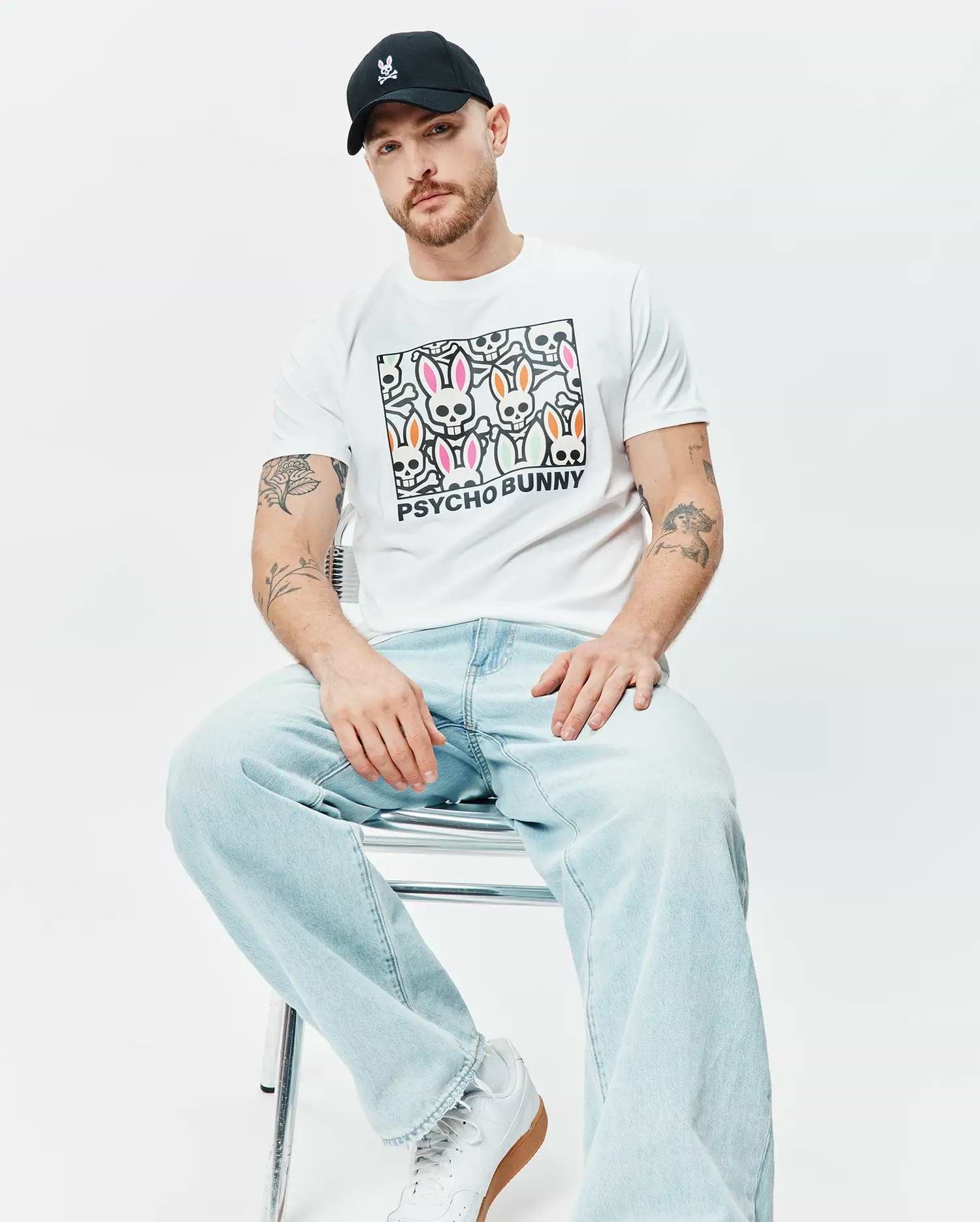 White Mens Byron Graphic Tee with a large central graphic print of colorful Psycho Bunny skull logos and PSYCHO BUNNY text, paired with a black cap featuring a small pink embroidered Psycho Bunny logo. (4)