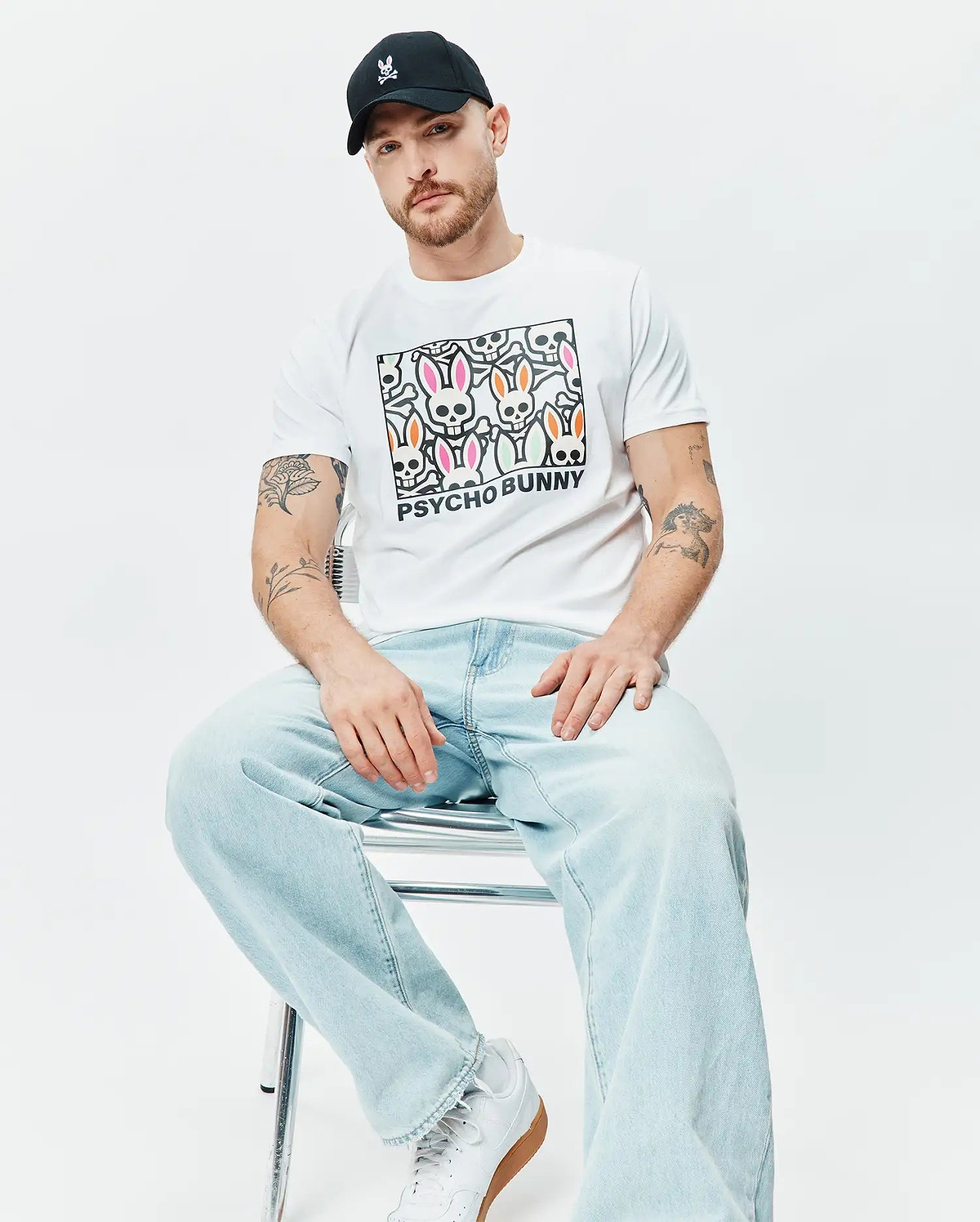 White Mens Byron Graphic Tee with a large central graphic print of colorful Psycho Bunny skull logos and PSYCHO BUNNY text, paired with a black cap featuring a small pink embroidered Psycho Bunny logo. (4)