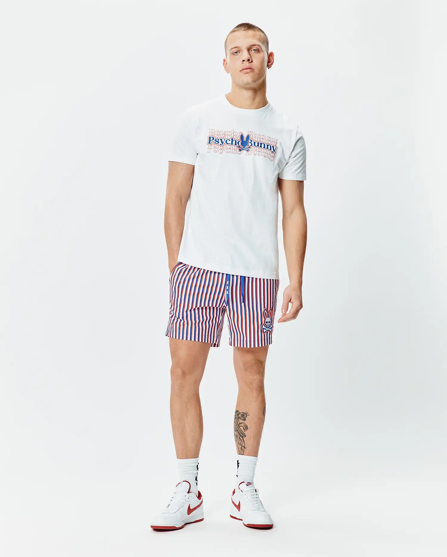White Mens John Back Graphic Tee with a large layered blue and red Psycho Bunny text and skull logo on the chest, paired with white, red, and blue striped shorts featuring a small embroidered bunny skull logo on the left leg.