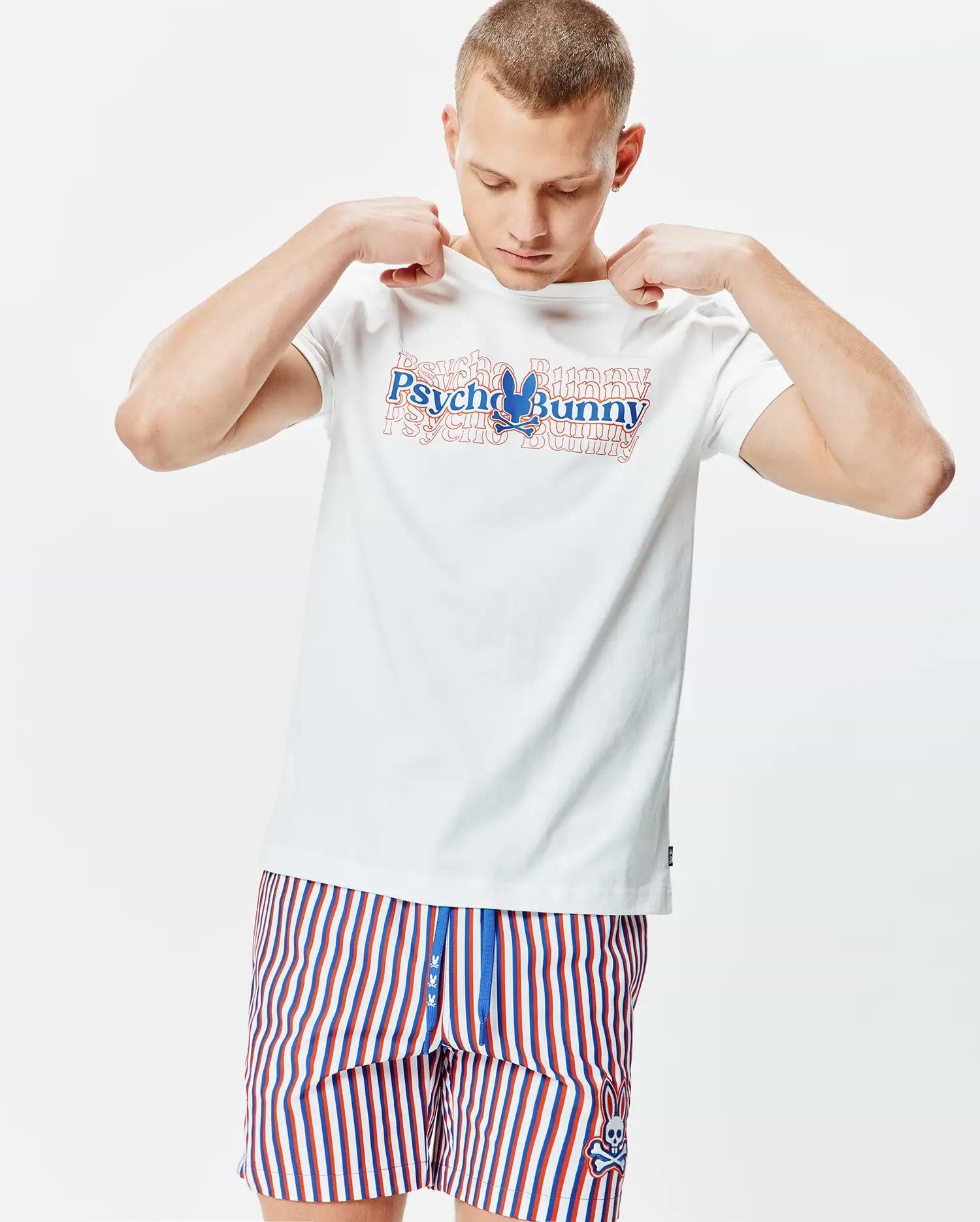 Mens John Back Graphic Tee, a white jersey t-shirt with a large layered red and blue Psycho Bunny chest graphic, paired with red, white, and blue striped shorts featuring a small embroidered bunny skull logo.