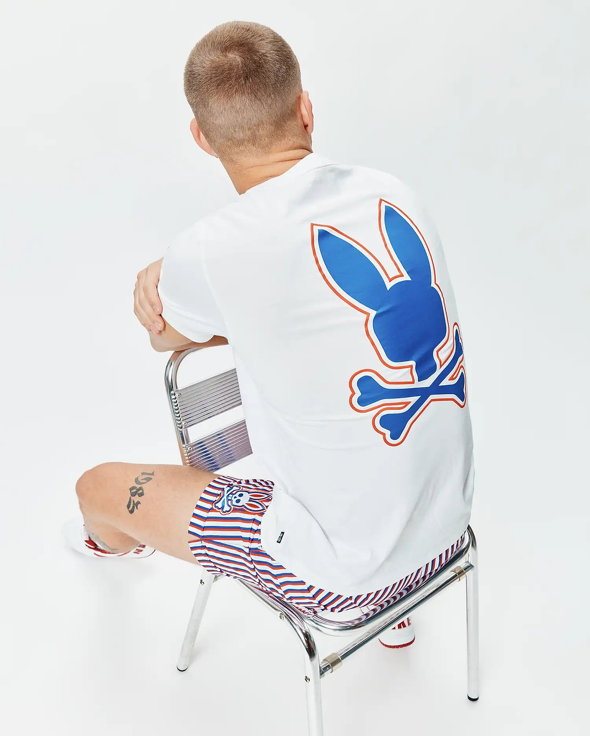 Mens John Back Graphic Tee, a white classic fit jersey t-shirt with a large blue and orange Psycho Bunny skull logo graphic on the back, paired with red, white, and blue striped shorts featuring a small bunny skull logo.