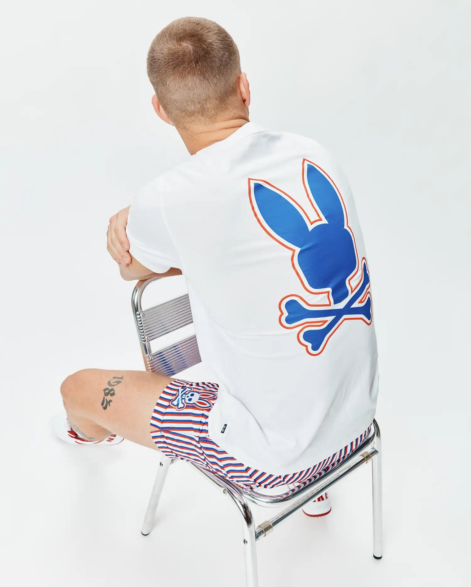 Mens John Back Graphic Tee, a white classic fit jersey t-shirt with a large blue and orange Psycho Bunny skull logo graphic on the back, paired with red, white, and blue striped shorts featuring a small bunny skull logo.