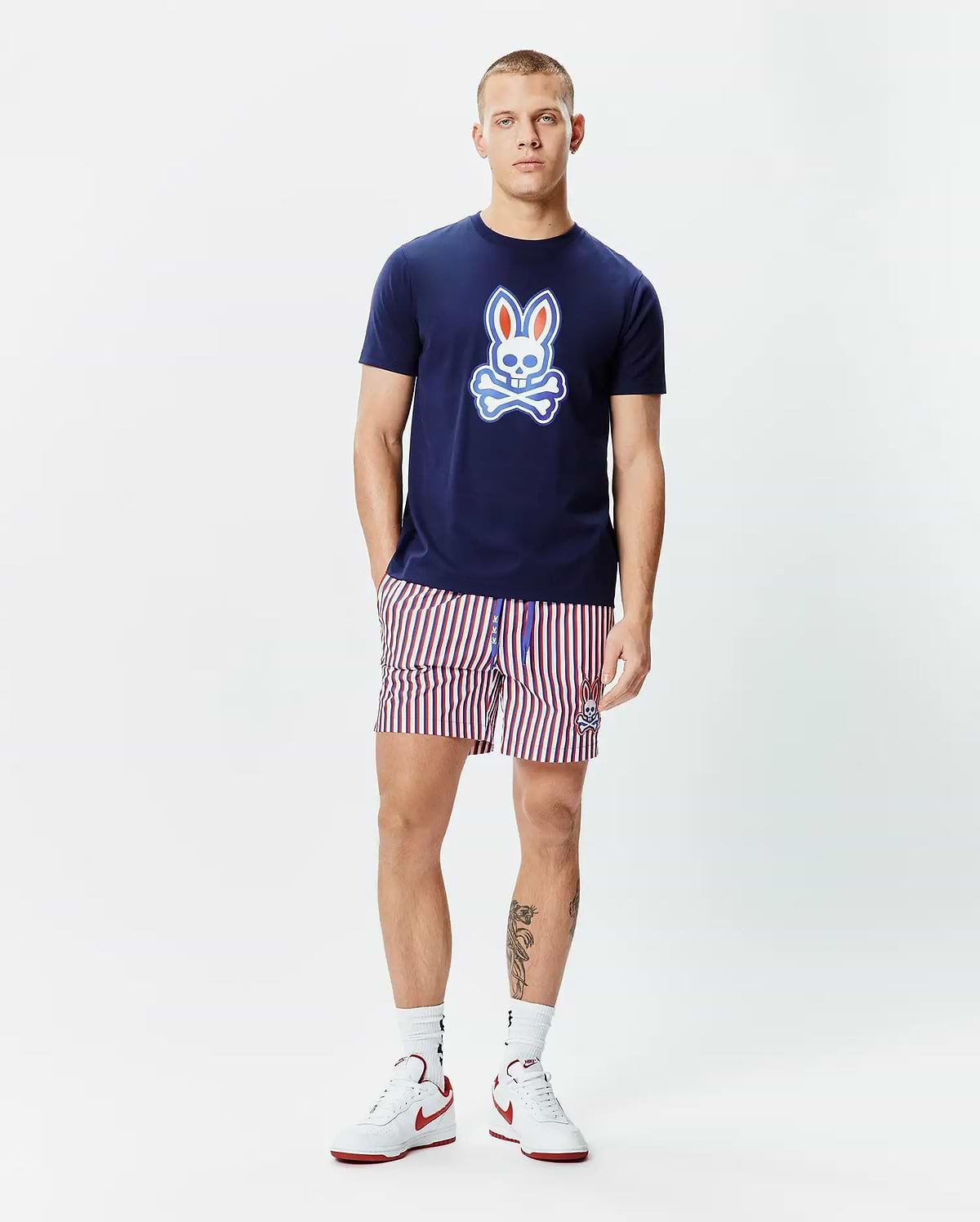 Mens John Graphic Tee, a navy blue jersey t-shirt with a large red, white, and light blue Psycho Bunny skull logo on the chest, paired with red, white, and navy striped shorts featuring a small embroidered bunny logo.