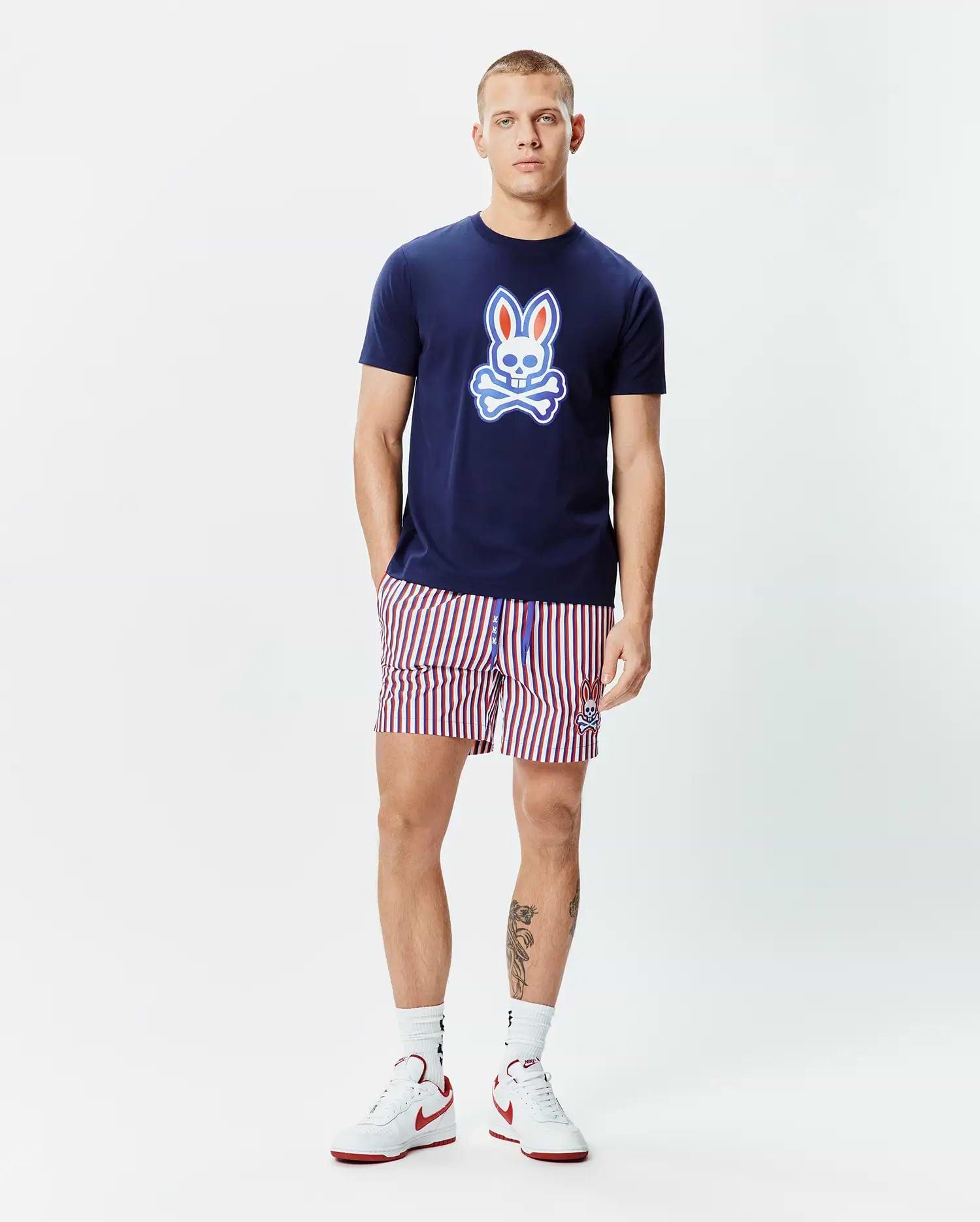 Mens John Graphic Tee, a navy blue jersey t-shirt with a large red, white, and light blue Psycho Bunny skull logo on the chest, paired with red, white, and navy striped shorts featuring a small embroidered bunny logo.