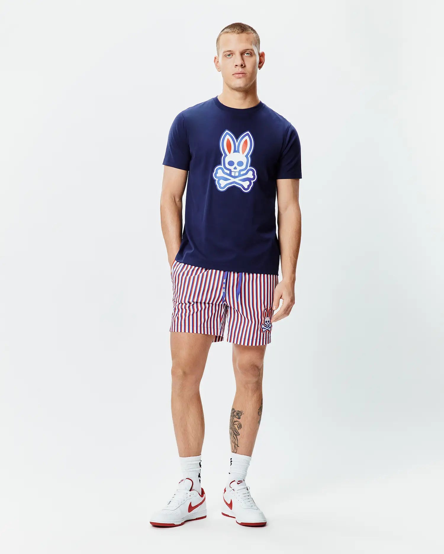 Mens John Graphic Tee, a navy blue jersey t-shirt with a large red, white, and light blue Psycho Bunny skull logo on the chest, paired with red, white, and navy striped shorts featuring a small embroidered bunny logo.