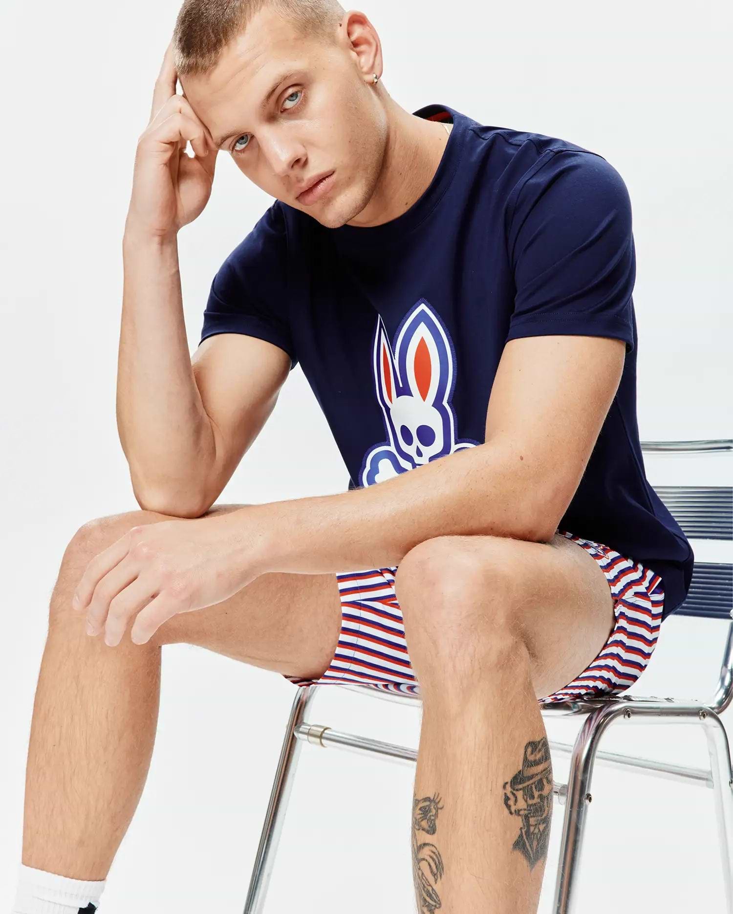 Mens John Graphic Tee, navy jersey, large white, red, blue Psycho Bunny skull logo on chest, paired with relaxed fit white, red, blue striped shorts.