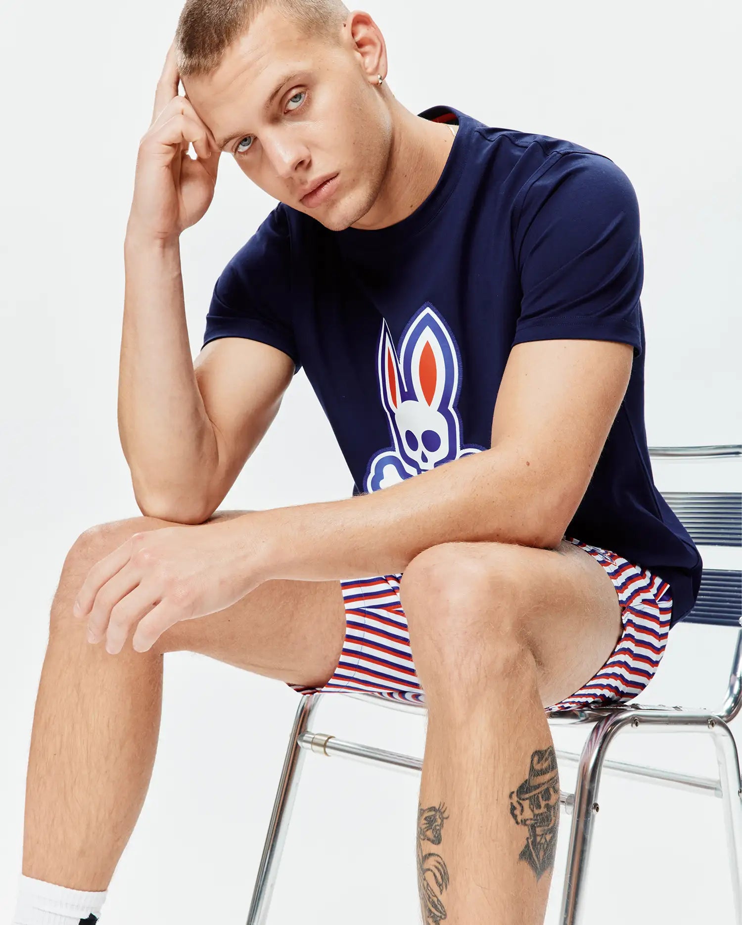 Mens John Graphic Tee, navy jersey, large white, red, blue Psycho Bunny skull logo on chest, paired with relaxed fit white, red, blue striped shorts.
