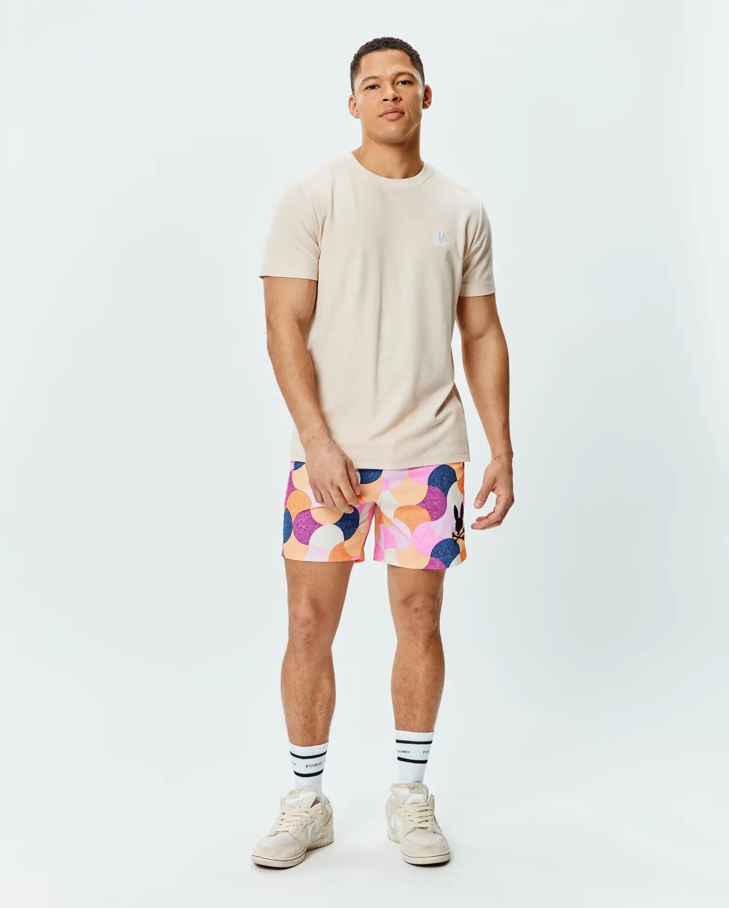 Mens Taylor Pique Tee in light beige with a small embroidered white Psycho Bunny logo on the left chest, paired with multi-color abstract patterned shorts featuring a black bunny logo on the right thigh.