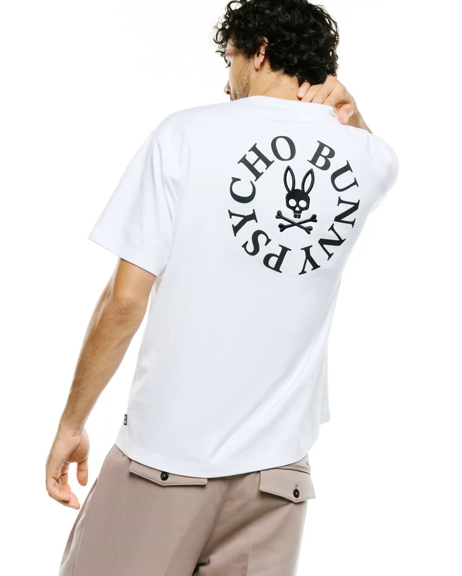 Mens Olek Relaxed Back Graphic Tee, a white relaxed fit jersey t-shirt featuring a large black circular Psycho Bunny skull logo and text graphic on the back, with a small woven tag on the side. (1)