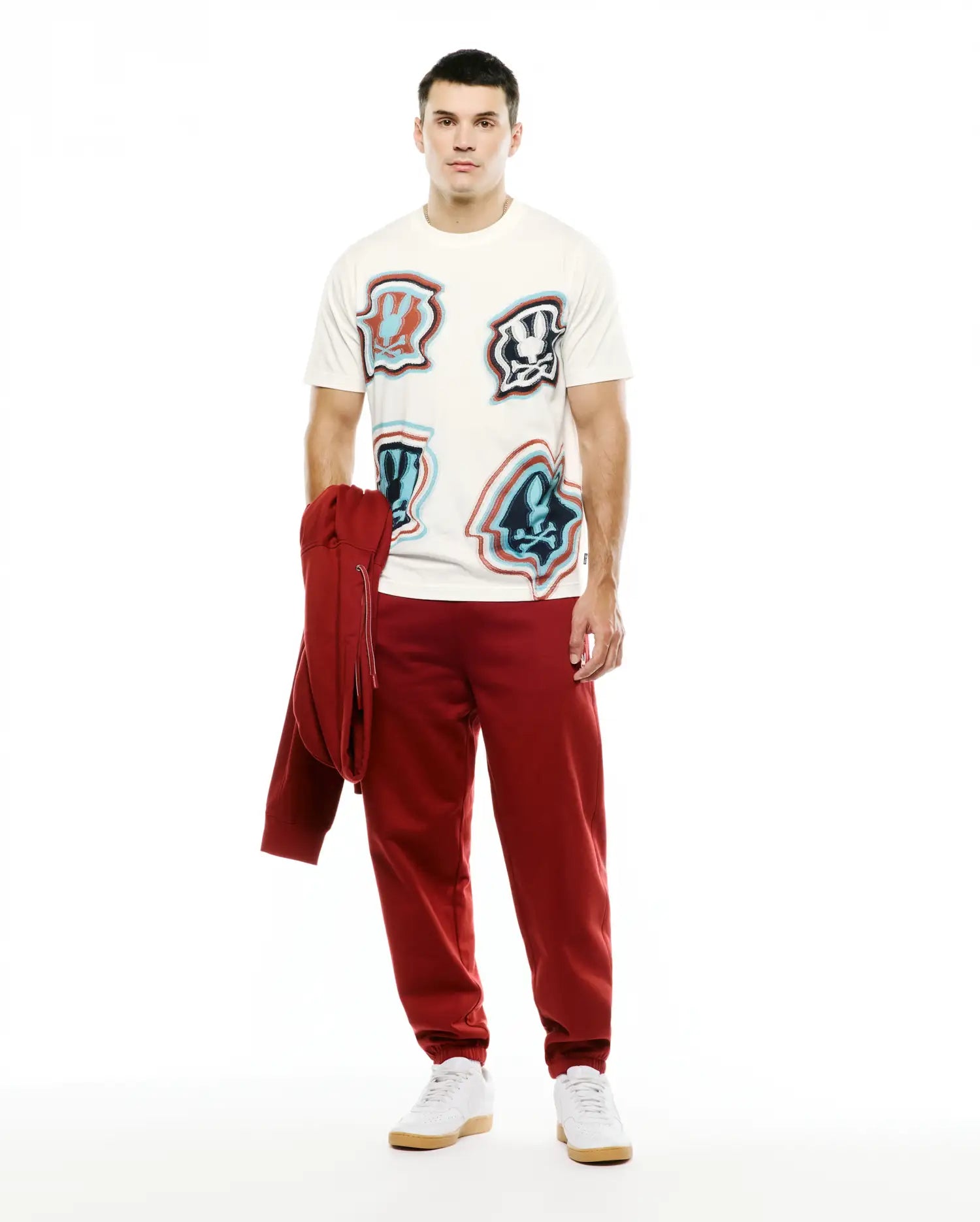Men's Klaus Jacquard Tee in cream, a relaxed fit jersey t-shirt with multiple iconic distorted red, light blue, and dark blue Psycho Bunny skull logos. Styled with relaxed deep red french terry sweatpants and a matching hoodie. (4)