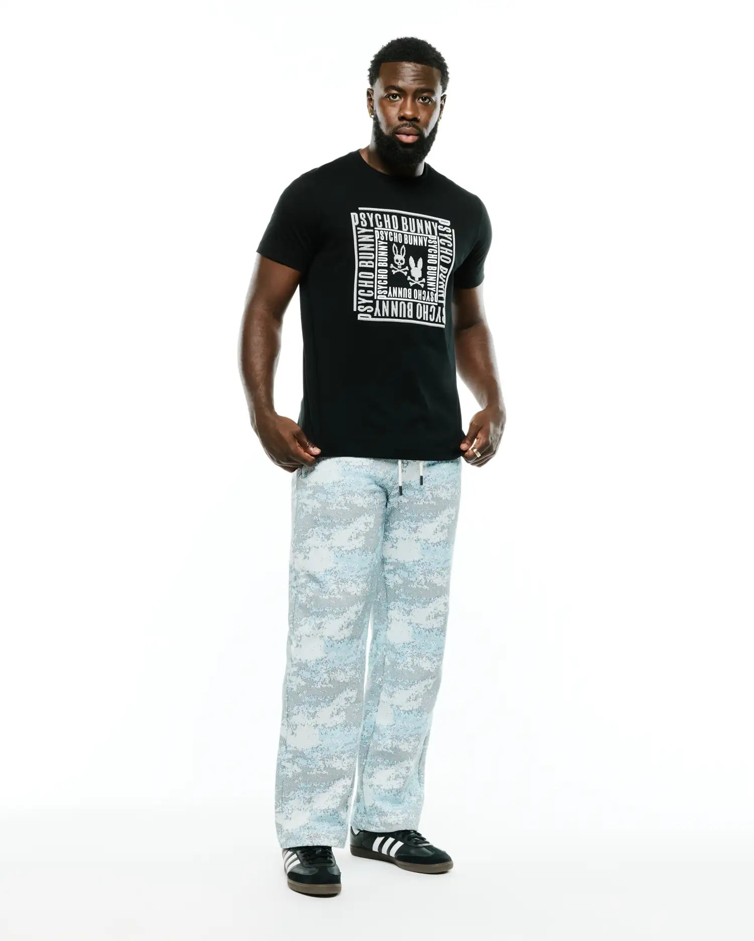 Black Men's Raj Wordmark Graphic Tee, classic fit, with a large white Psycho Bunny repeating wordmark and skull logo chest graphic. Paired with relaxed light blue and grey digital camo pants. (5)