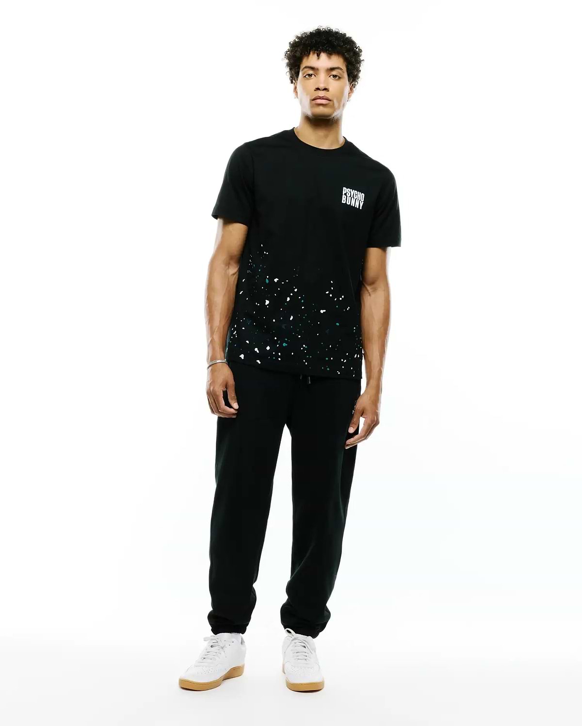 Mens Malcolm Graphic Tee. Black classic fit jersey tee with white and teal paint splatter graphic on the lower front and a small white Psycho Bunny text logo on the left chest.