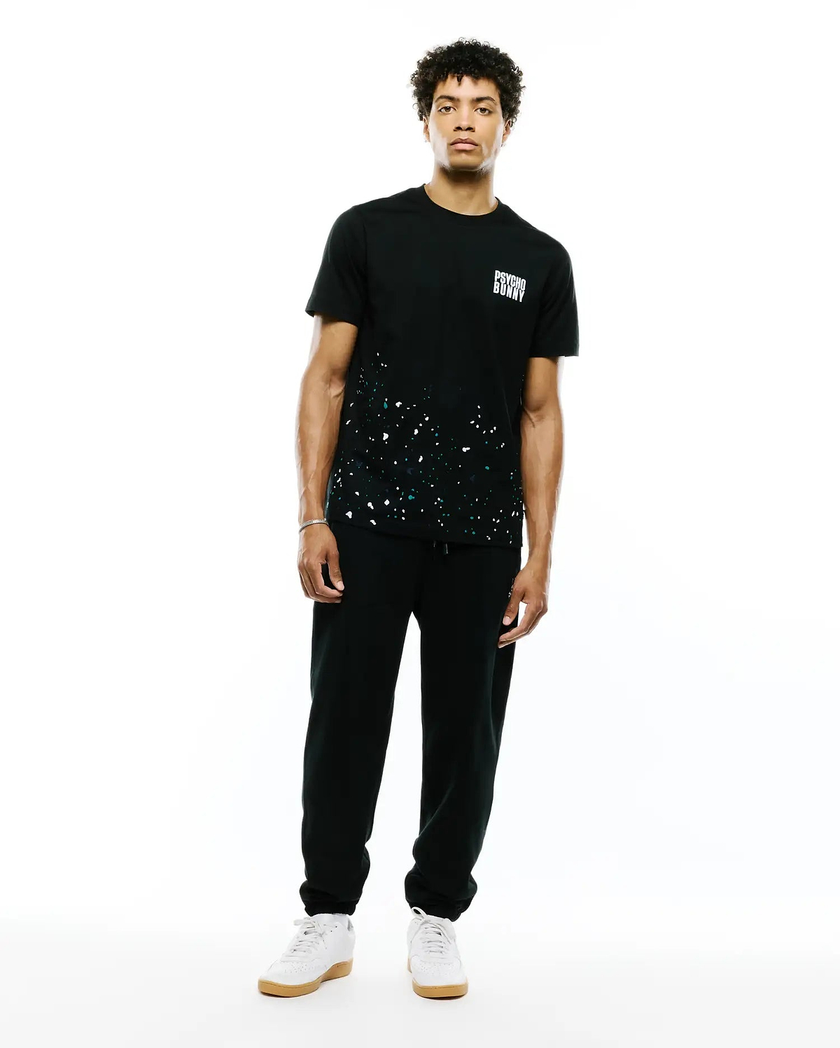 Mens Malcolm Graphic Tee. Black classic fit jersey tee with white and teal paint splatter graphic on the lower front and a small white Psycho Bunny text logo on the left chest.