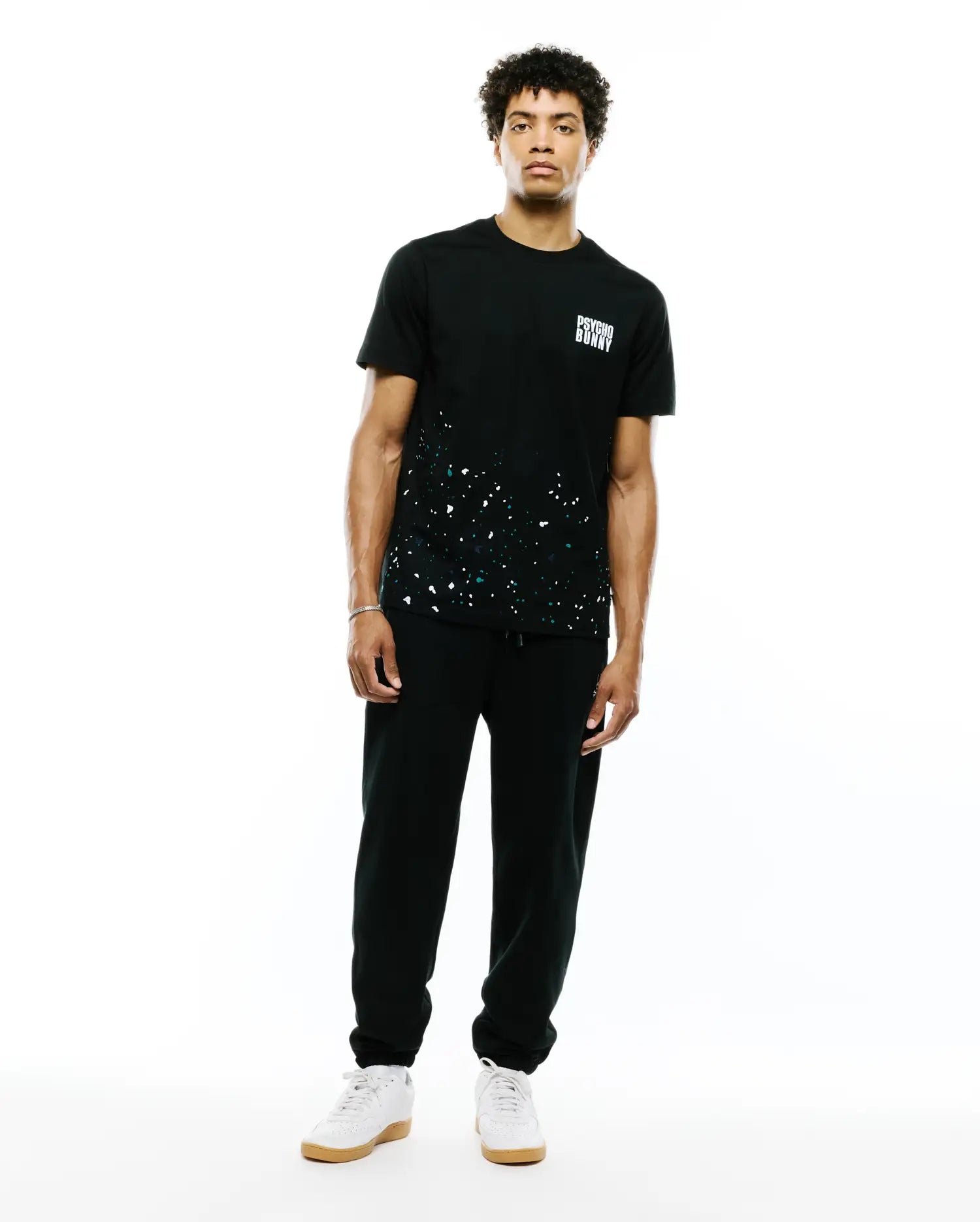 Mens Malcolm Graphic Tee. Black classic fit jersey tee with white and teal paint splatter graphic on the lower front and a small white Psycho Bunny text logo on the left chest.