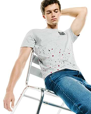 Mens Malcolm Graphic Tee, a classic fit heather grey jersey t-shirt featuring a small black Psycho Bunny text logo on the upper left chest and a vibrant red, black, and pink paint splatter graphic across the lower front.