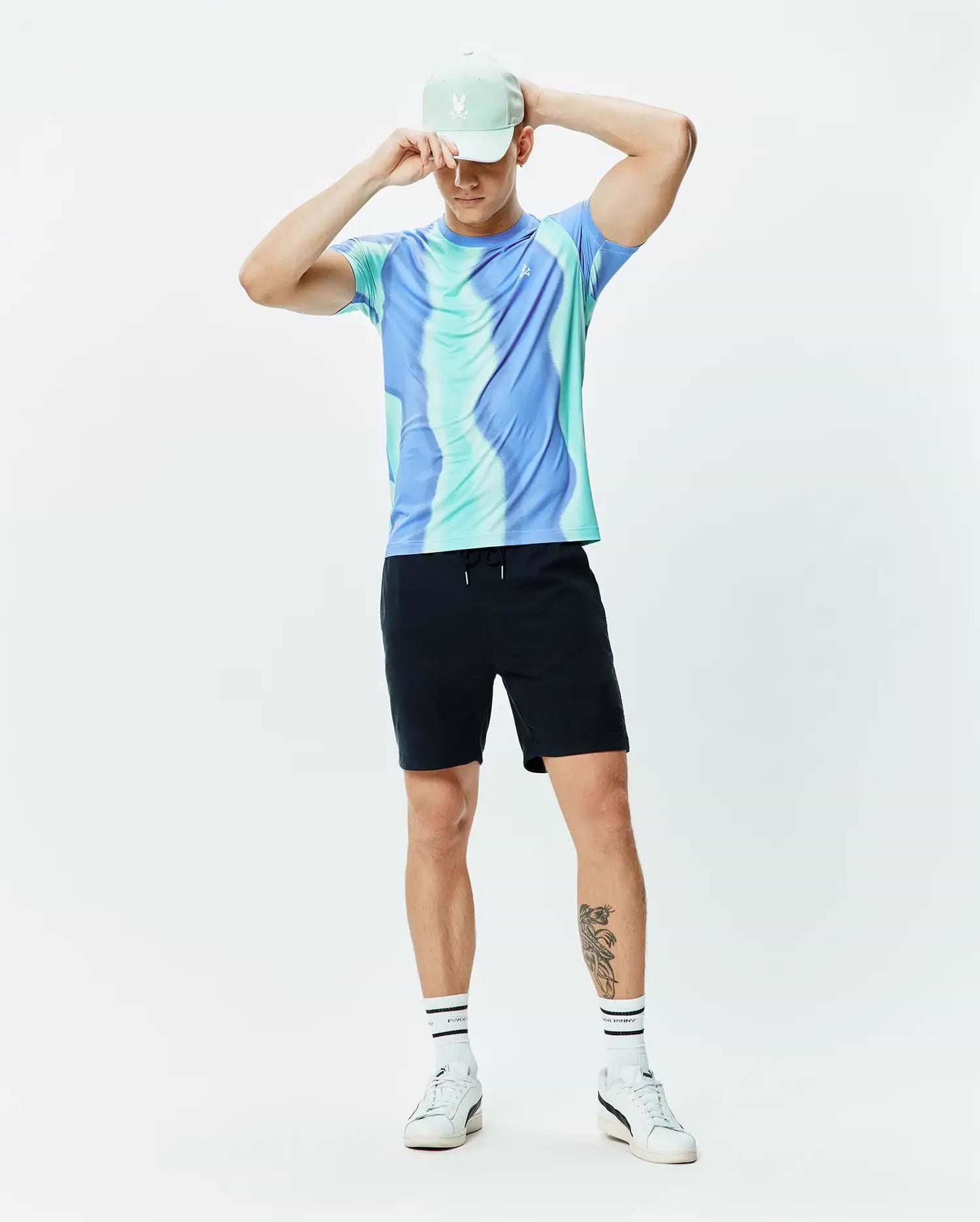 Mens Blaze Sport Tee in blue and teal swirl pattern with a small white Psycho Bunny logo on the chest, paired with black drawstring shorts and a mint green cap featuring an embroidered white bunny skull.