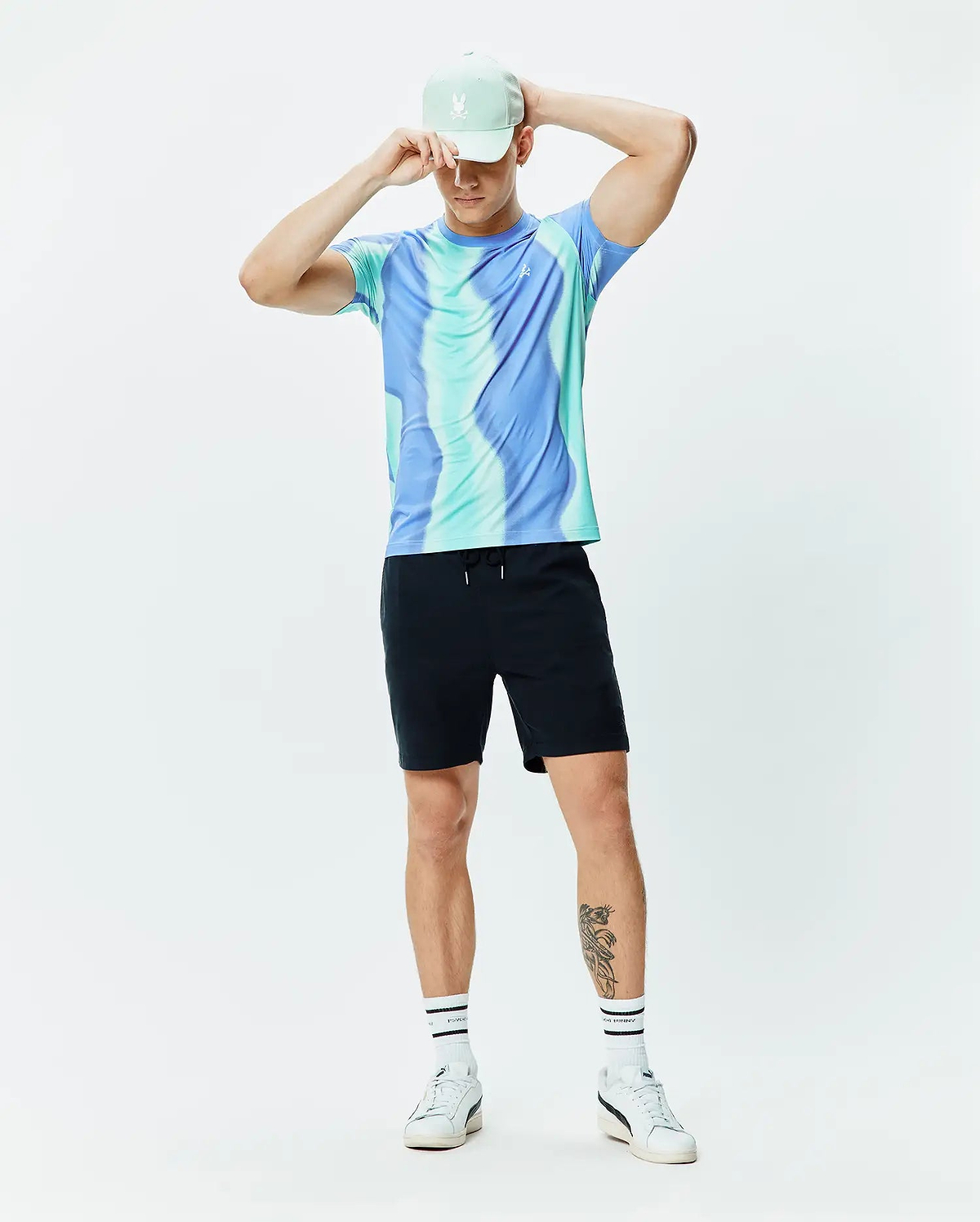 Mens Blaze Sport Tee in blue and teal swirl pattern with a small white Psycho Bunny logo on the chest, paired with black drawstring shorts and a mint green cap featuring an embroidered white bunny skull.