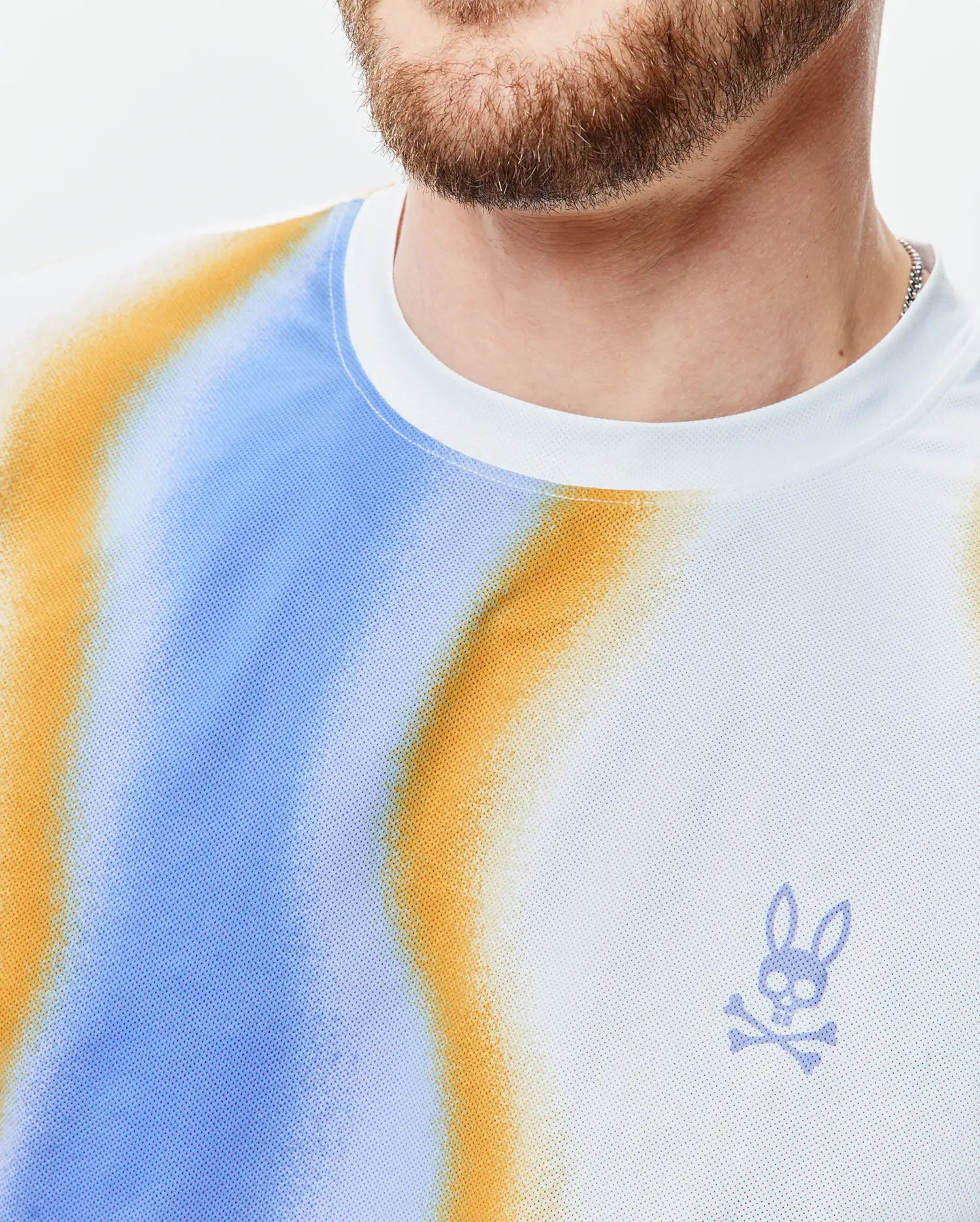 Mens Blaze Sport Tee, white performance mesh with blue and orange gradient pattern, featuring a small embroidered light purple Psycho Bunny skull logo on the left chest.