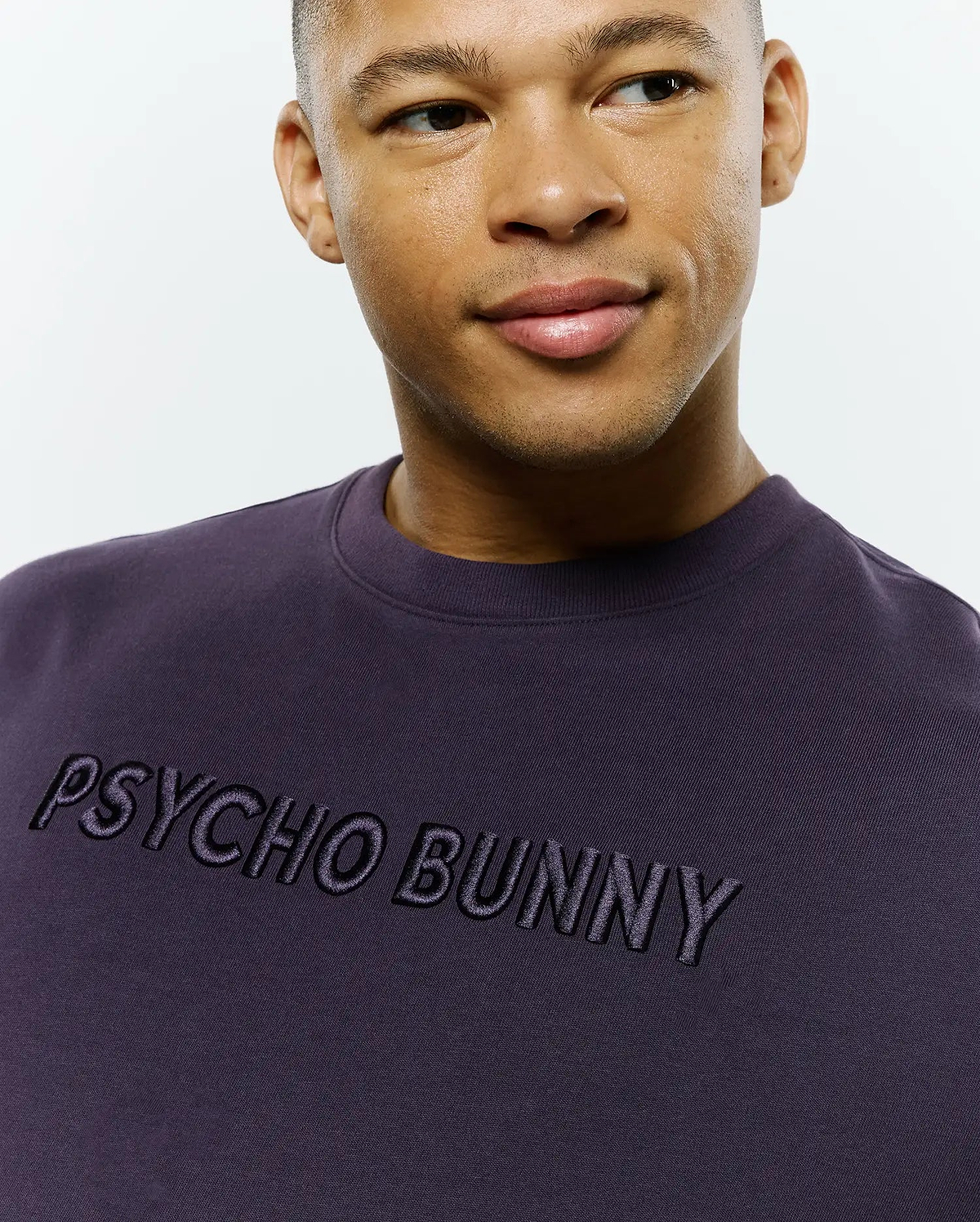 Dark purple Mens Windcrest Relaxed Fit Tee, crafted from smooth jersey knit, featuring a prominent black embroidered Psycho Bunny text logo across the chest.