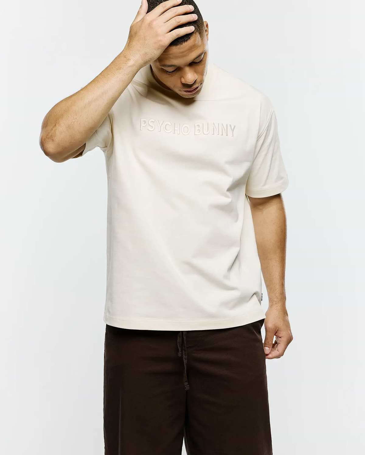 Mens Windcrest Relaxed Fit Tee in cream smooth jersey, featuring a large tonal embossed PSYCHO BUNNY text logo centered on the chest and a small black side seam tag. (3)