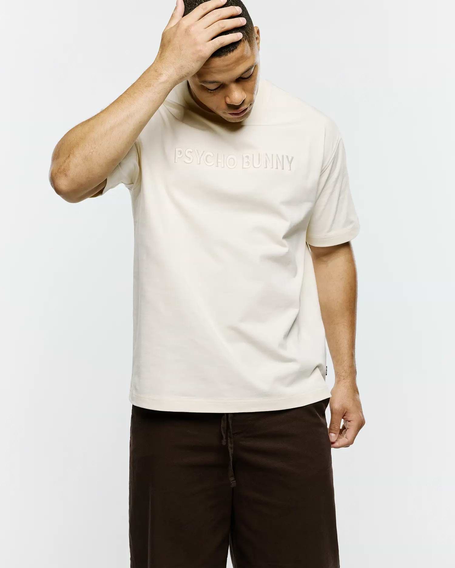 Mens Windcrest Relaxed Fit Tee in cream smooth jersey, featuring a large tonal embossed PSYCHO BUNNY text logo centered on the chest and a small black side seam tag. (3)