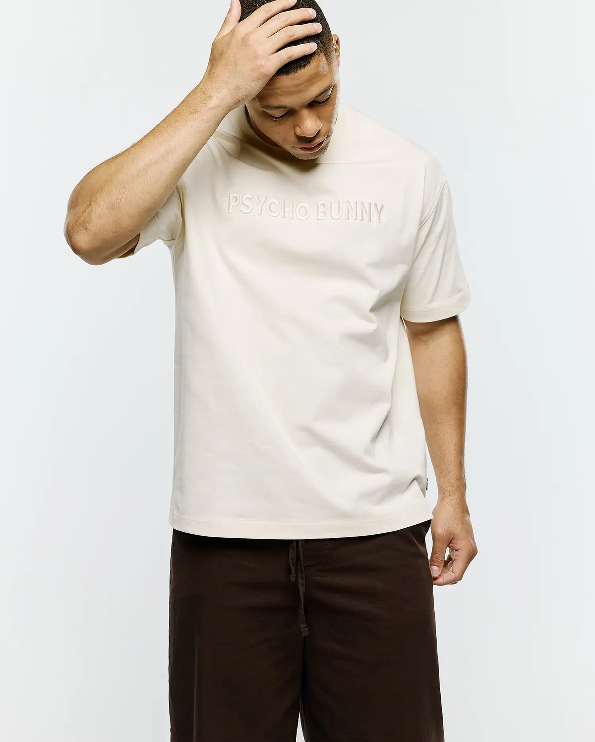 Mens Windcrest Relaxed Fit Tee in cream smooth jersey, featuring a large tonal embossed PSYCHO BUNNY text logo centered on the chest and a small black side seam tag. (3)