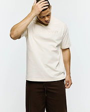 Mens Windcrest Relaxed Fit Tee in cream smooth jersey, featuring a large tonal embossed PSYCHO BUNNY text logo centered on the chest and a small black side seam tag. (3)