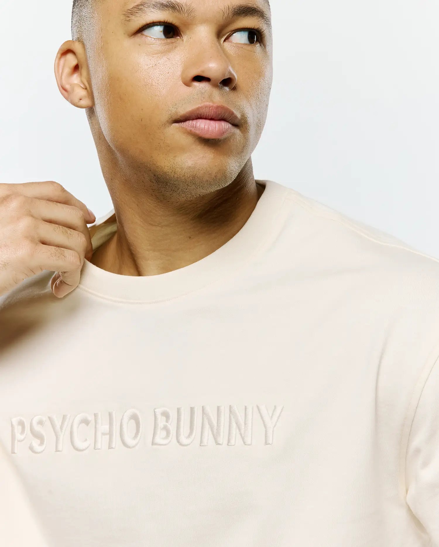 Mens Windcrest Relaxed Fit Tee in a luxurious cream soft jersey knit, showcasing a prominent, tonal embroidered PSYCHO BUNNY text across the chest. (2)
