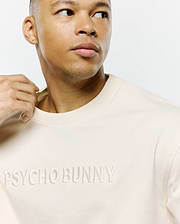 Mens Windcrest Relaxed Fit Tee in a luxurious cream soft jersey knit, showcasing a prominent, tonal embroidered PSYCHO BUNNY text across the chest. (2)