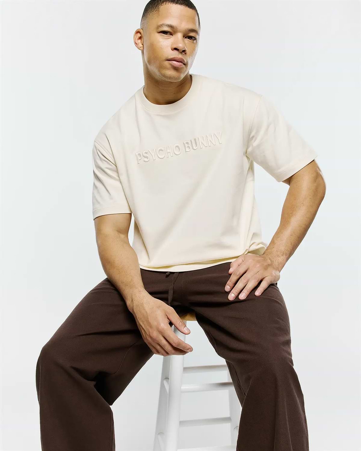 Mens Windcrest Relaxed Fit Tee in cream, featuring a smooth jersey fabric and a large, tonal embroidered Psycho Bunny text logo on the center chest. (1)