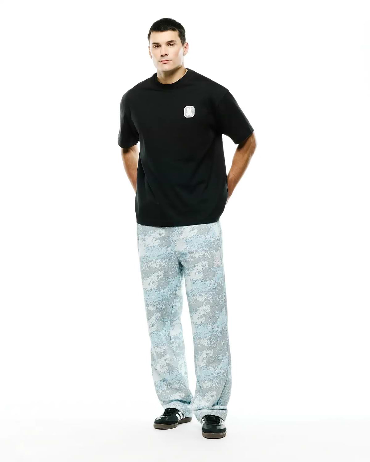 Men's Samson Oversized Bunny Tee in black jersey knit, featuring a small white embroidered Psycho Bunny skull logo on the left chest, paired with light blue and grey digital camouflage relaxed-fit pants. (4)