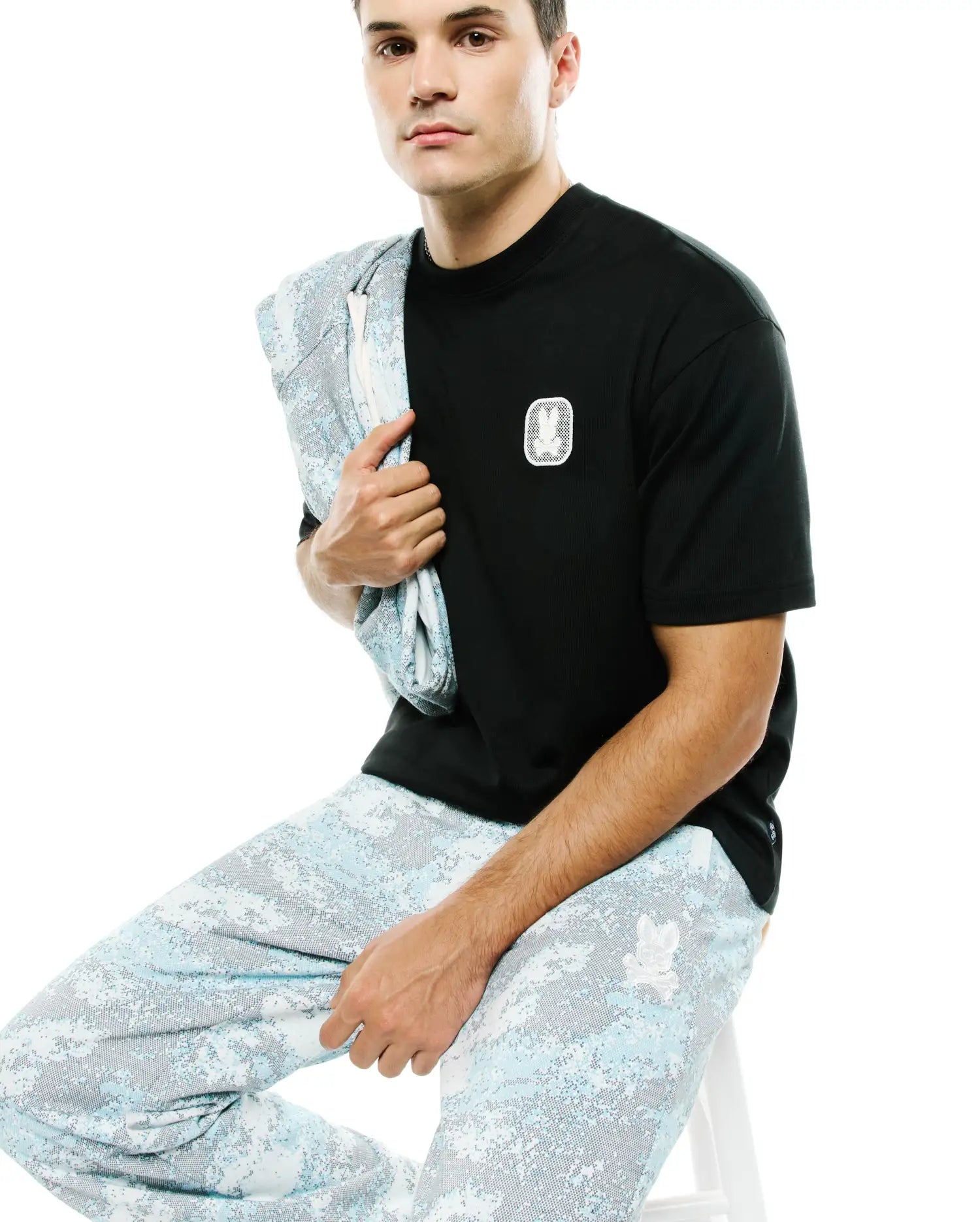 Men's Samson Oversized Bunny Tee: Black ribbed t-shirt with an oversized fit, featuring a small white embroidered Psycho Bunny skull logo on the left chest. Complemented by light blue digital camo pants. (3)